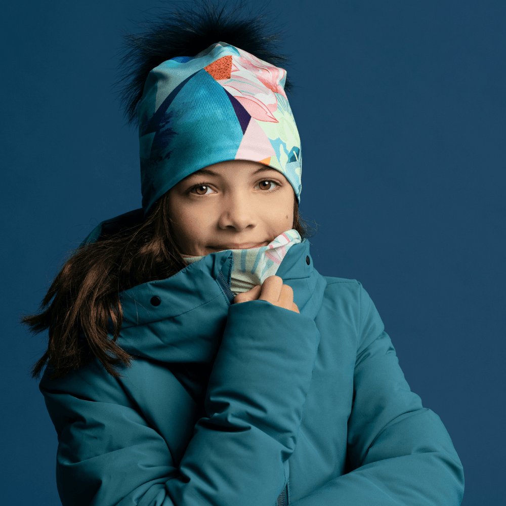 Close-up of a young girl wearing a patterned beanie and neck warmer, smiling against a blue background.