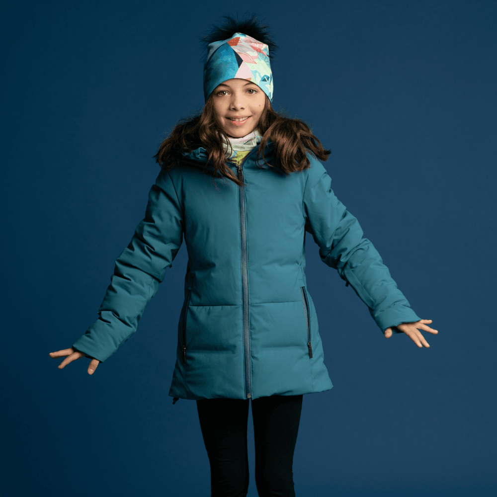 A young girl wearing a teal winter jacket, a matching patterned beanie, and a neck warmer, smiling.