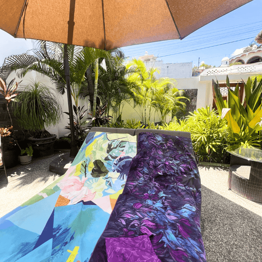 Two large microfiber towels, one with abstract floral art and the other with a purple floral design, spread out on lounge chairs under an umbrella.
