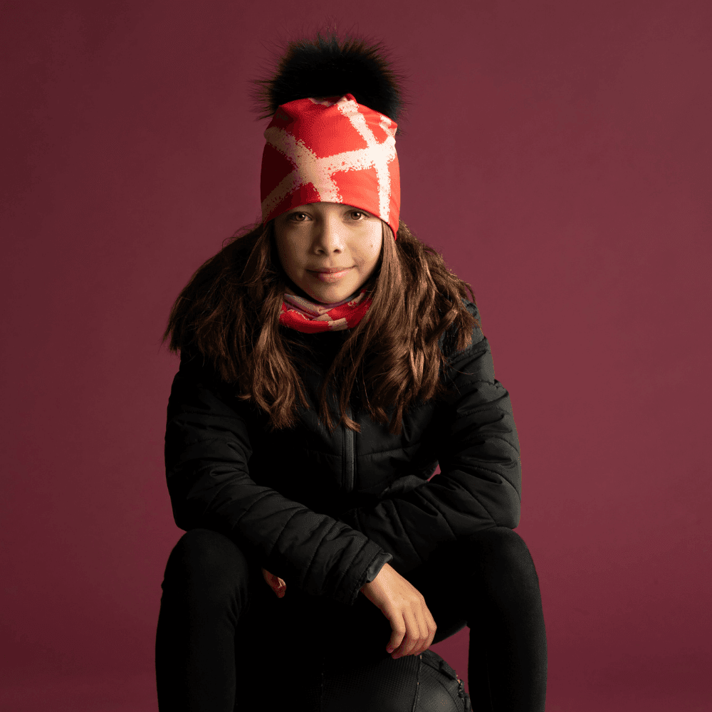 Young girl wearing the Peachy-Red Beanie and a matching neck tube, sitting in front of a burgundy backdrop.