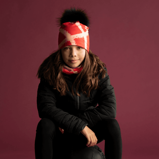 Young girl wearing the Peachy-Red Beanie and a matching neck tube, sitting in front of a burgundy backdrop.