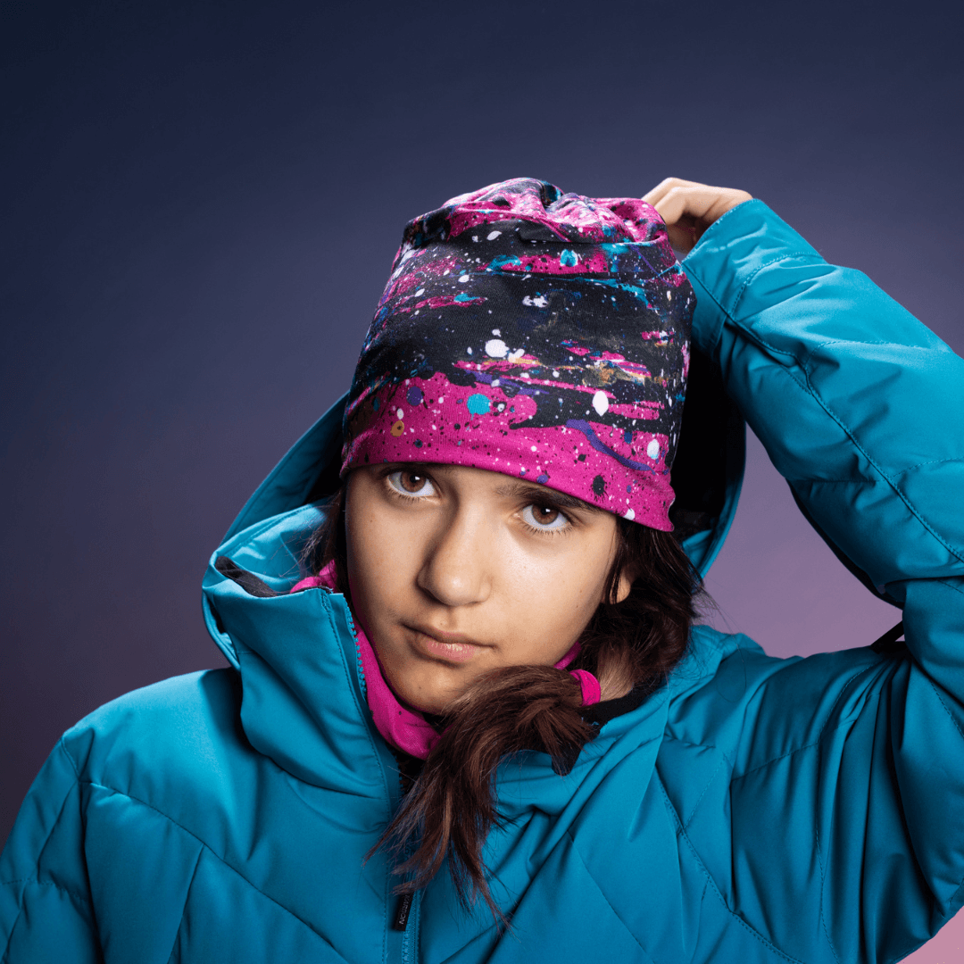 Young girl in a blue ski jacket wearing a pink abstract print beanie and pink neck tube, looking at the camera.