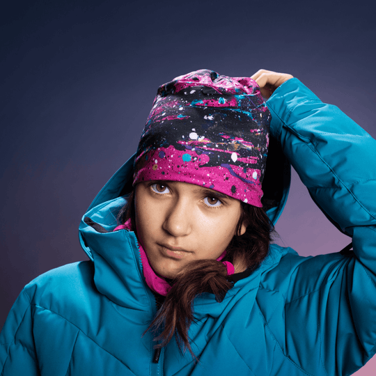 Young girl in a blue ski jacket wearing a pink abstract print beanie and pink neck tube, looking at the camera.