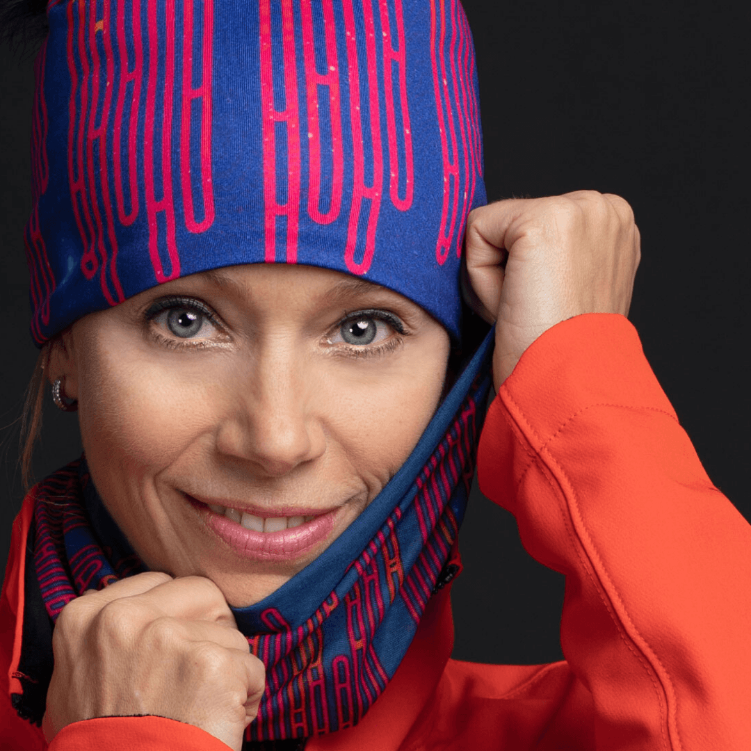 A woman smiles, pulling on a blue and red patterned beanie and showing off a matching neck gaiter, wearing an orange jacket.