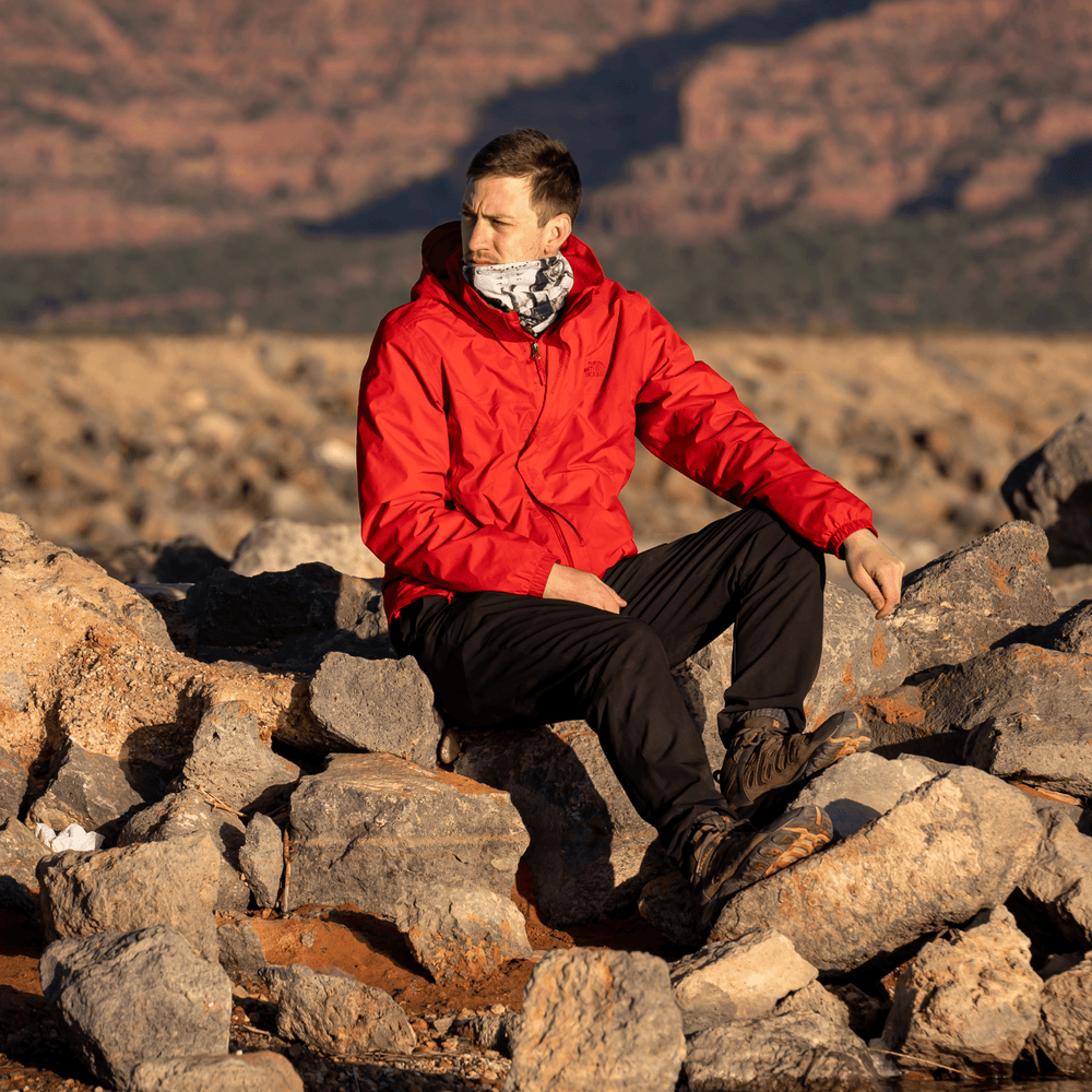 Man in a red jacket sitting on rocks outdoors, wearing the black and white patterned tube as a neck gaiter.