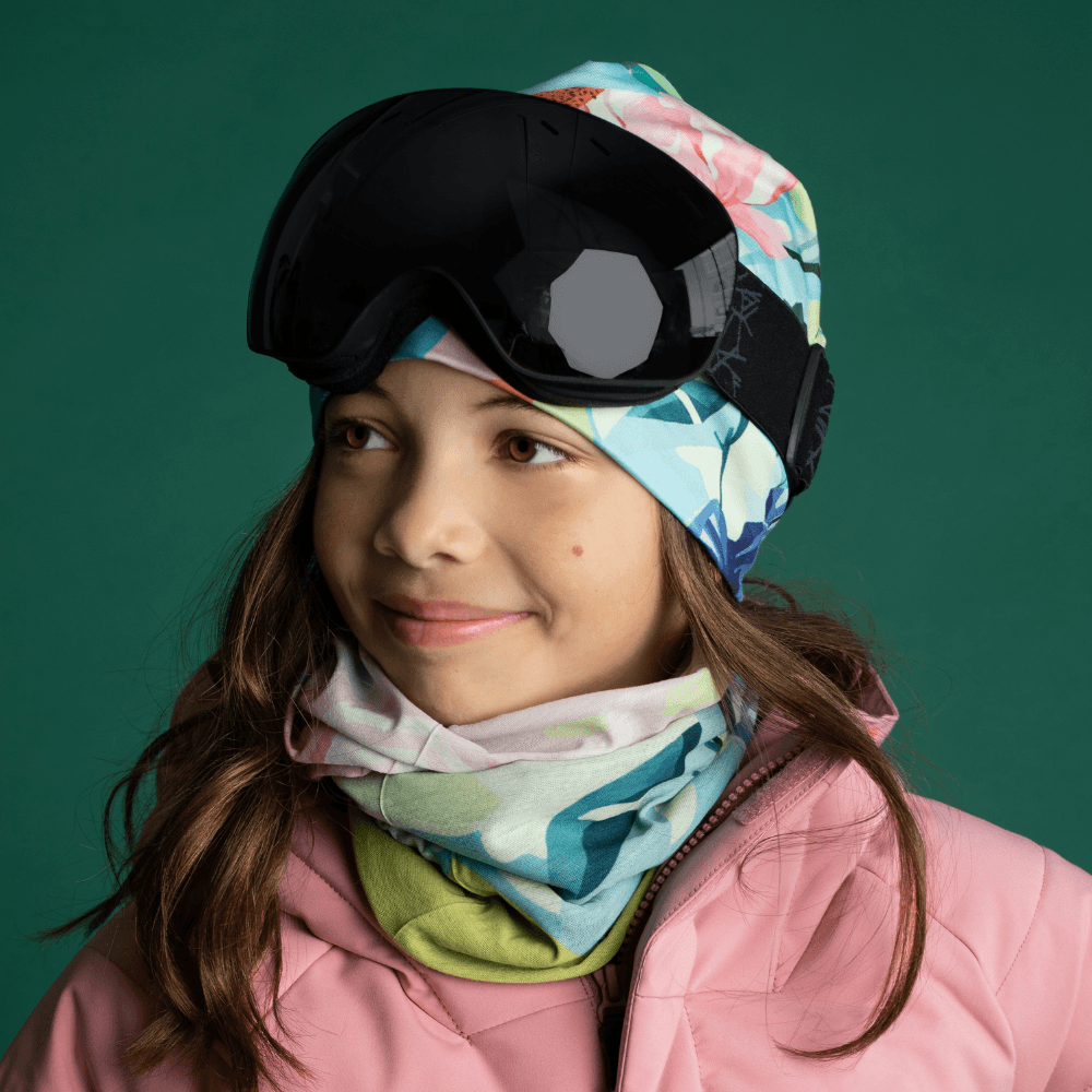A young girl wearing a pink ski jacket and goggles, with the floral neck tube and matching beanie.