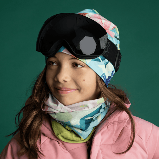 A young girl wearing a pink ski jacket and goggles, with the floral neck tube and matching beanie.