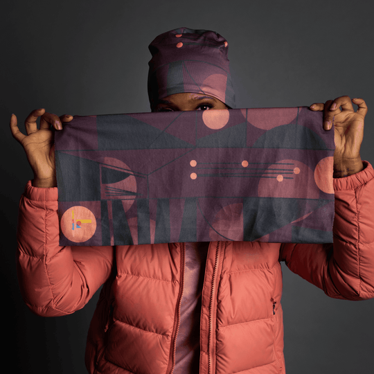 A person wearing a coral puffer jacket holds up a rectangular fabric tube with a geometric purple and dark grey pattern, their face partially obscured.