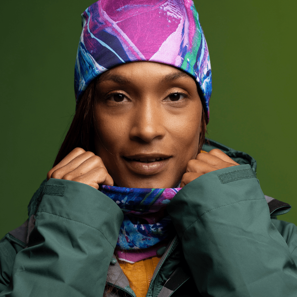 A woman wearing a green winter jacket, pulls up the collar to reveal a colorful, art-printed neck gaiter that matches the beanie she is wearing.