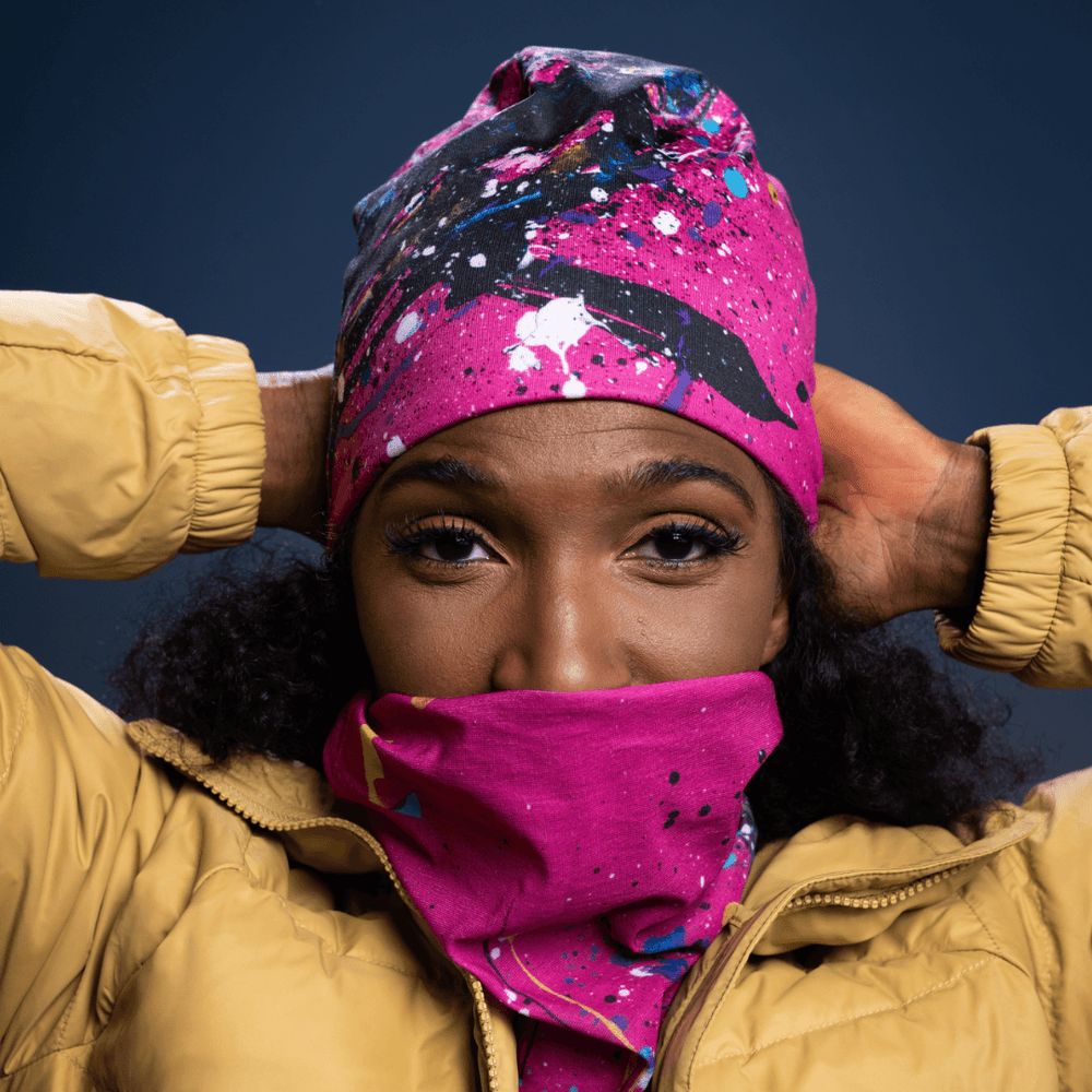 Woman wearing a yellow puffer jacket pulls a pink, blue, and black abstract paint splatter tube up over her face.