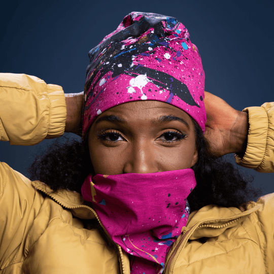 Woman wearing a yellow puffer jacket pulls a pink, blue, and black abstract paint splatter tube up over her face.