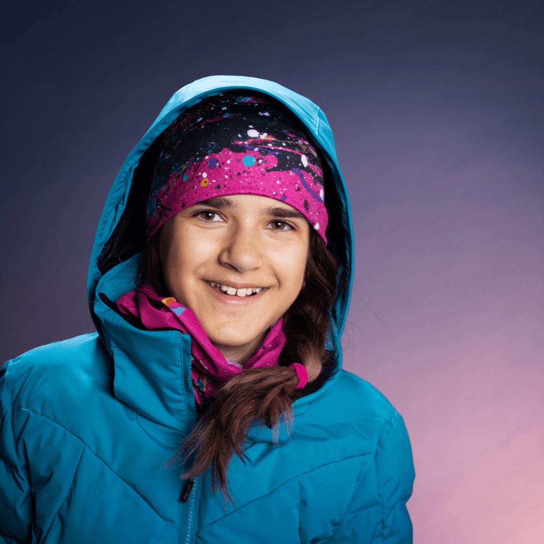 Young girl in a blue hooded winter jacket smiles, wearing a pink, blue, and black abstract paint splatter headscarf.