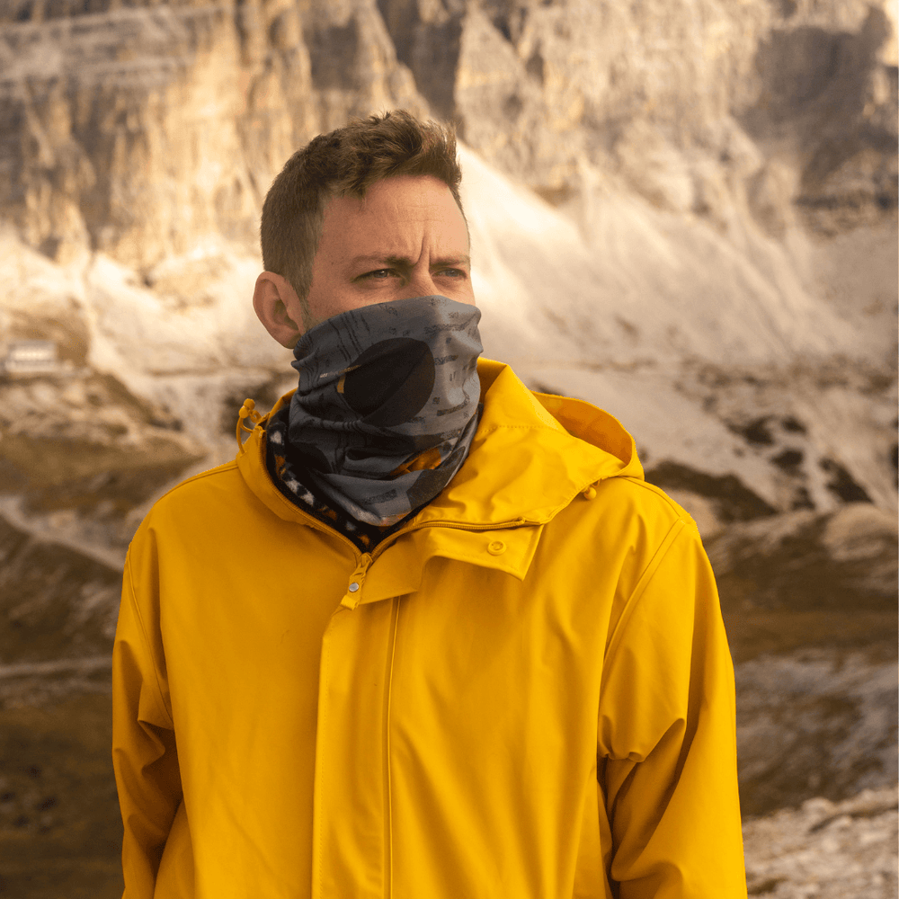 Man wearing a yellow raincoat and a neck gaiter with a dark grey and black abstract print covering his face in a snowy mountain landscape.