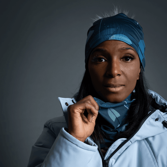 A woman wearing a light blue winter coat pulls the patterned POLAR Umbrella Teal neck tube higher around her neck, revealing the artistic design.