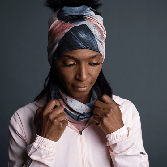 Woman wearing a light pink jacket, with the POLAR Vie en Rose Multifunction Tube styled as a headband and neck warmer, revealing a matching beanie with a pompom.