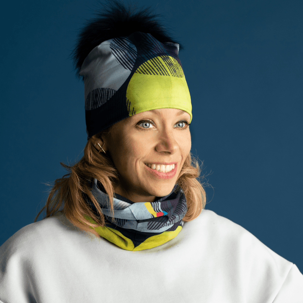 Woman smiling, wearing a colorful beanie and a matching neck warmer, with a blue background.