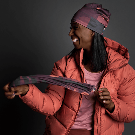 Woman in a coral puffer jacket holding a Purple Geometrics neck tube, smiling.