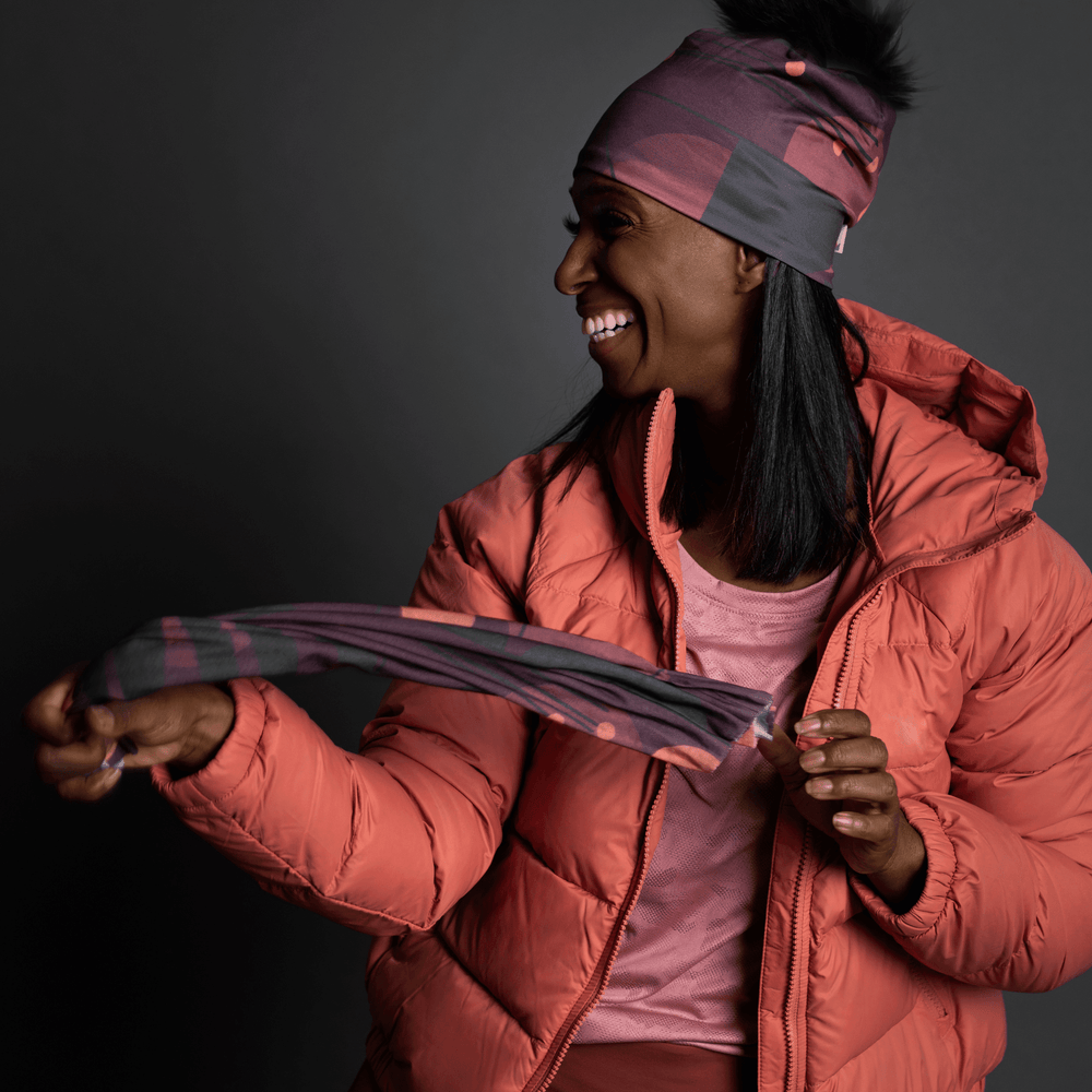 Woman in a coral puffer jacket holds a matching Purple Geometrics neck gaiter, smiling.