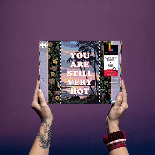 Hands holding the "Puzzle 500 pieces «Still Very Hot »" box against a purple background.