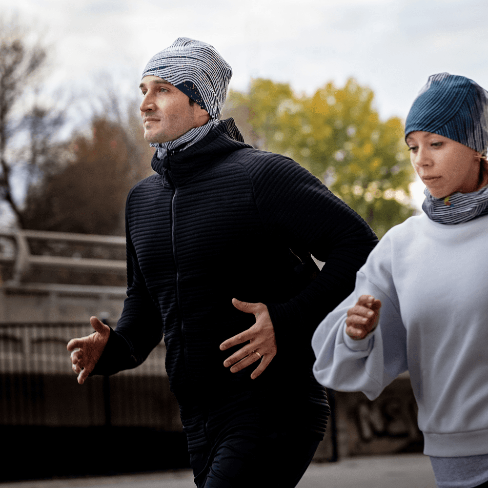 Man and woman wearing matching Dark Steel print beanies and neck gaiters while jogging outdoors in a city park.