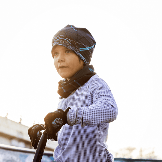 Young boy wearing a beanie and neck warmer with the Robocats print, standing on a scooter in a skatepark.