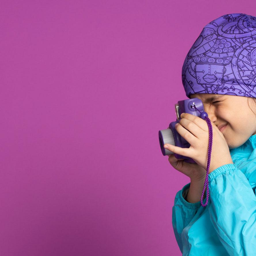 A young girl wearing the purple robot art beanie and a blue jacket, holding a purple toy camera.