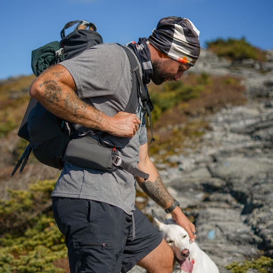 Man wearing a black beanie and neck gaiter with abstract art, hiking on a rocky trail with a dog.