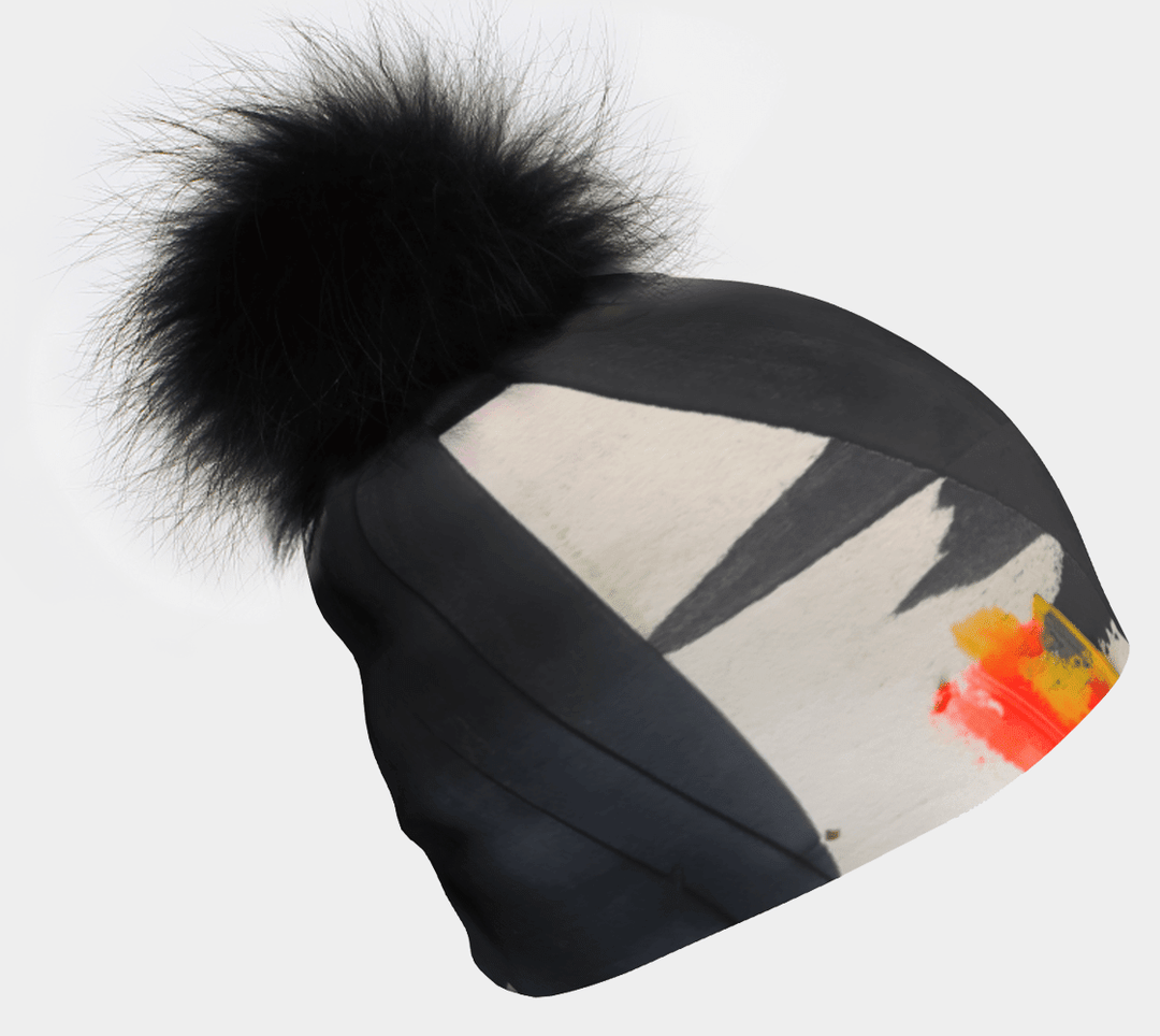 Close-up of a black beanie with abstract monochrome and vibrant orange and yellow art, topped with a large fluffy black pom-pom.