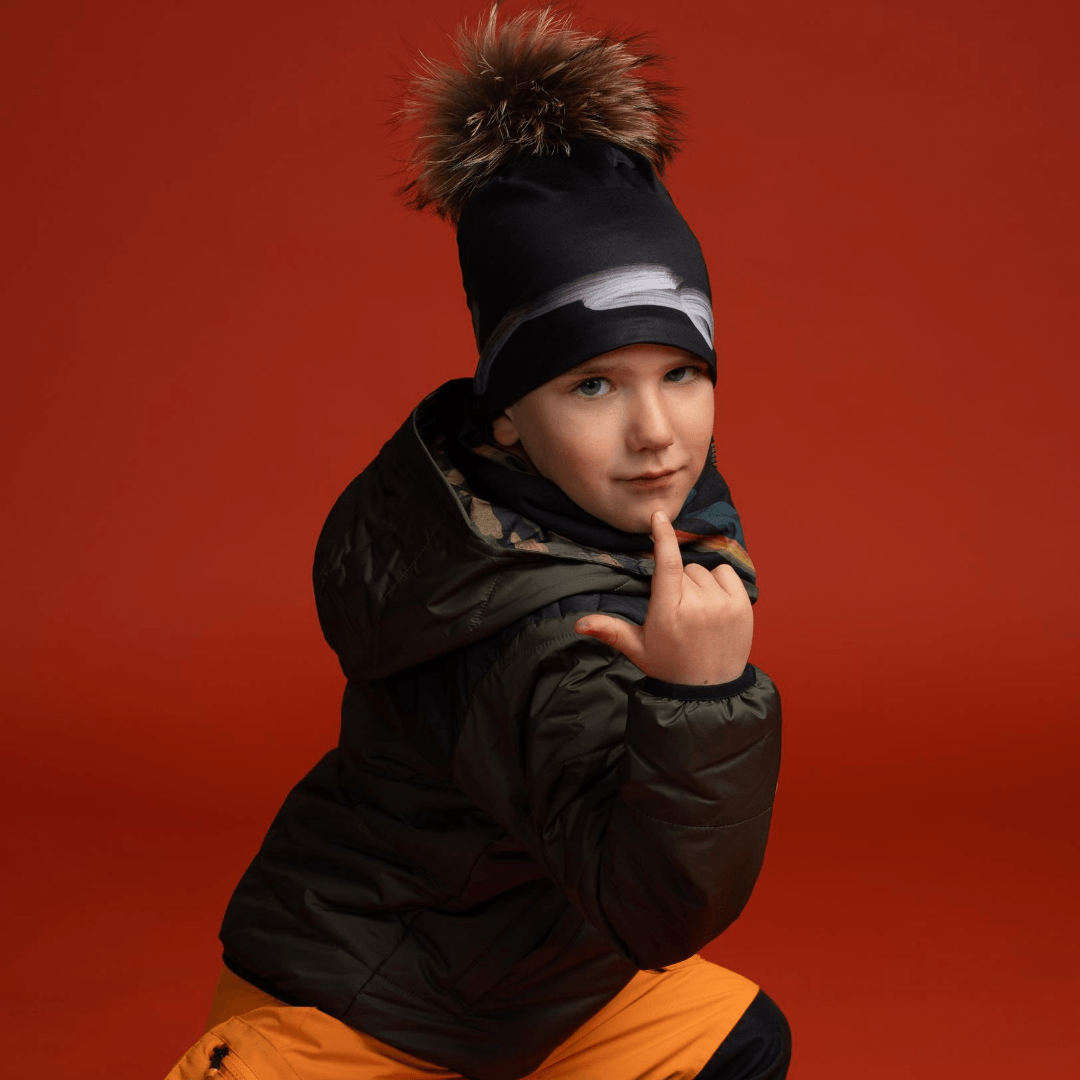 A young boy wearing the Signature Black Beanie with pompom and a winter jacket, posing against a red background.
