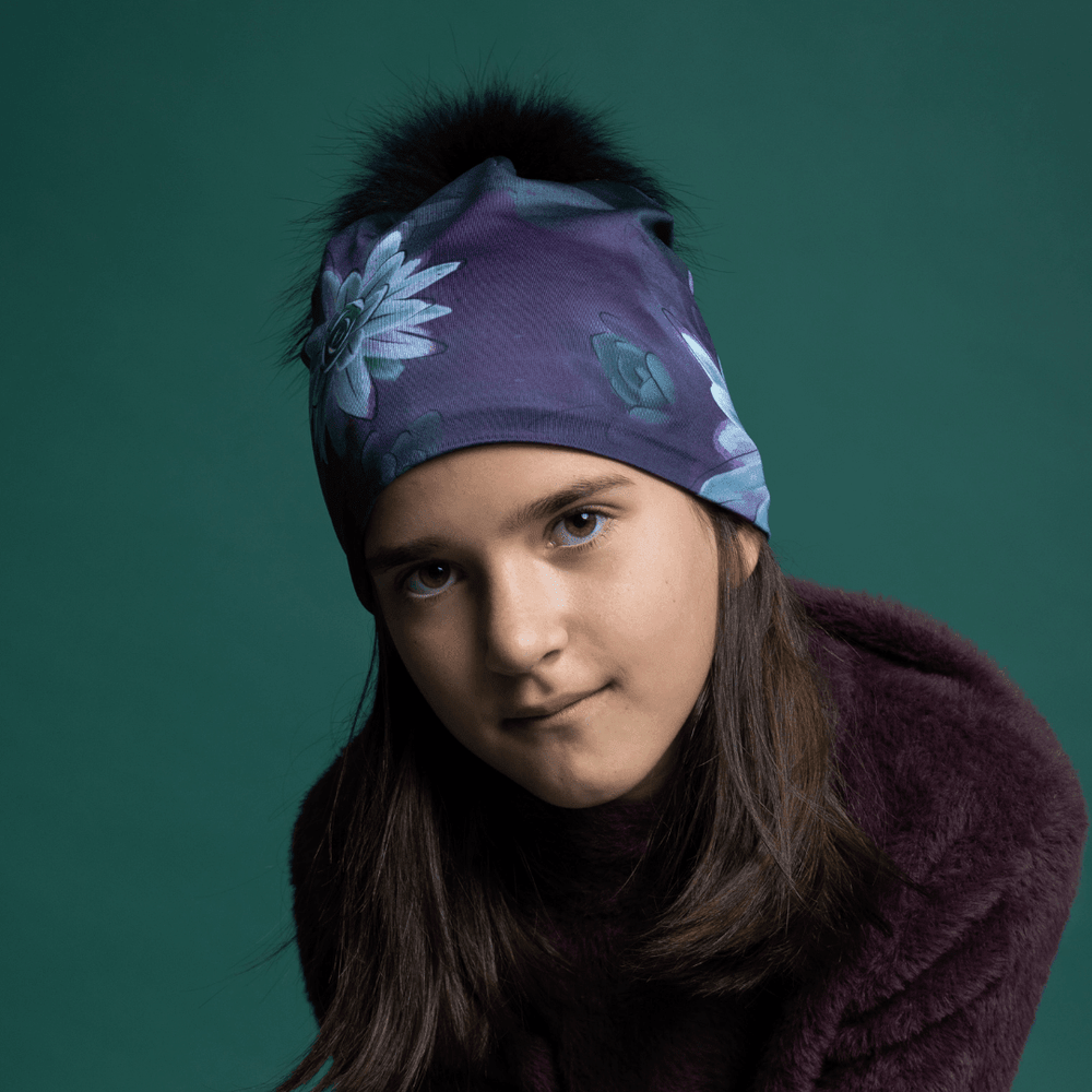 Young woman wearing a purple succulent-print beanie with black fur pompom, styled with a plum faux-fur coat against teal background