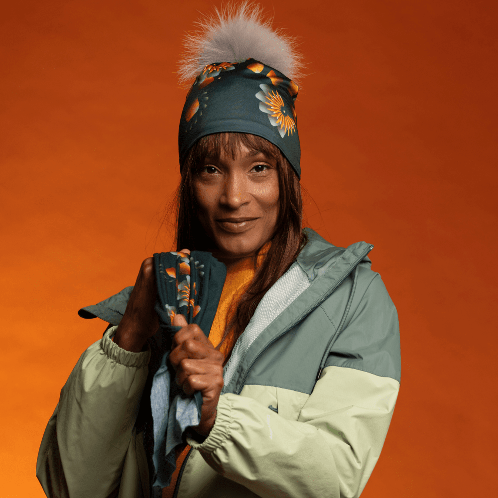 Woman wearing a dark green beanie with a green and orange floral pattern, holding a matching neck tube.
