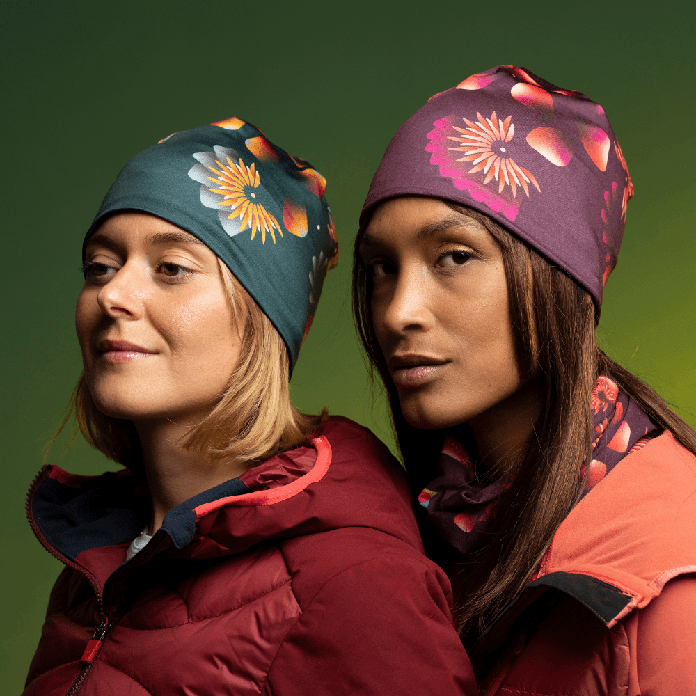 Two women wearing beanies with floral prints, one dark green and one purple, against a green background.