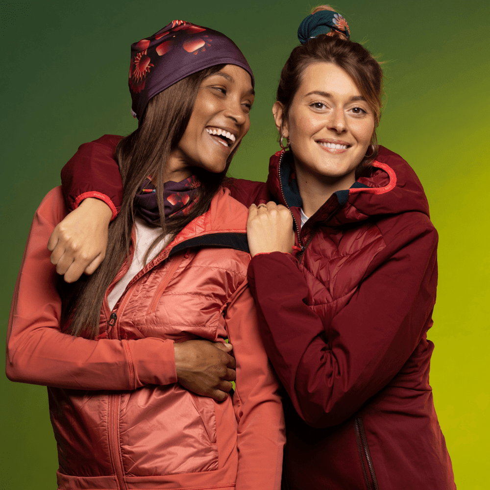 Deux femmes en manteaux d'hiver et tuques sourient, l'une portant une tuque marine et l'autre une tuque et un cache-cou à motifs.