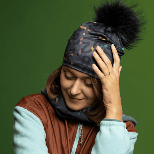 Woman wearing a gray beanie with a black pompom and matching neck gaiter, looking down thoughtfully.