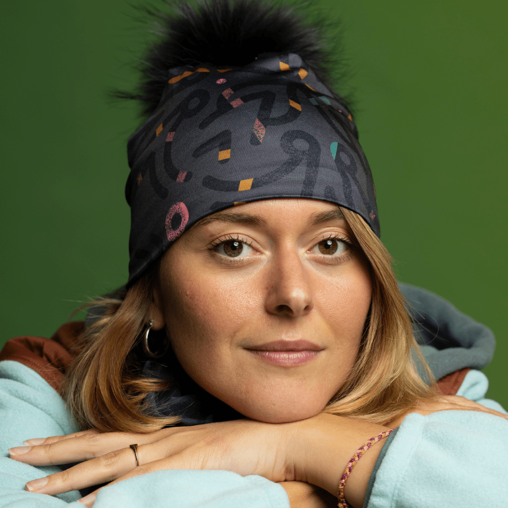 Woman wearing a gray beanie with a black pompom and matching neck gaiter, posing with hands clasped.