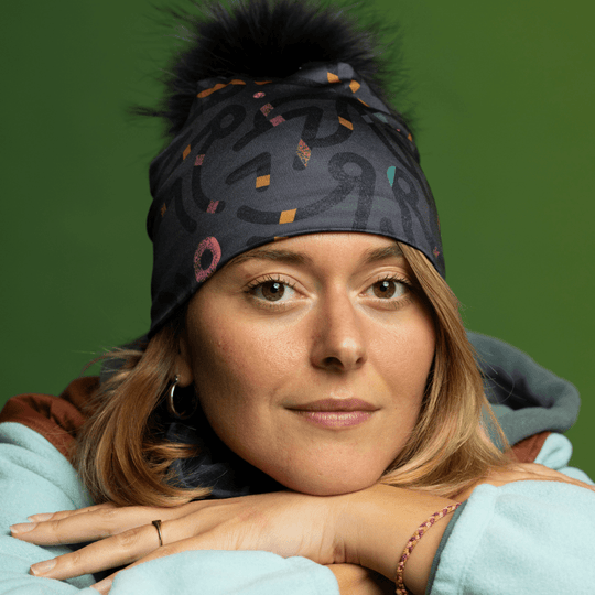 Woman wearing a gray beanie with a black pompom and matching neck gaiter, posing with hands clasped.