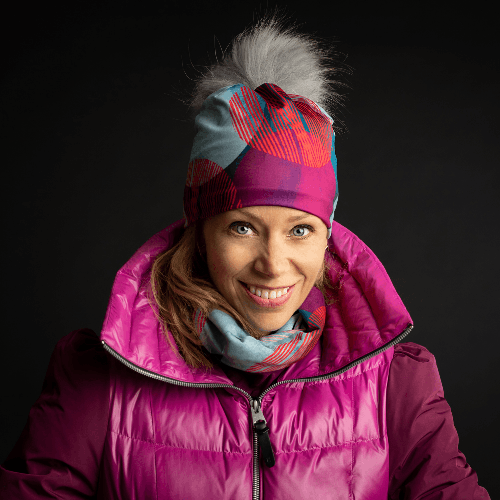 Woman smiling, wearing the Teal-Dots Beanie and a matching neck warmer, layered under a bright pink winter jacket.