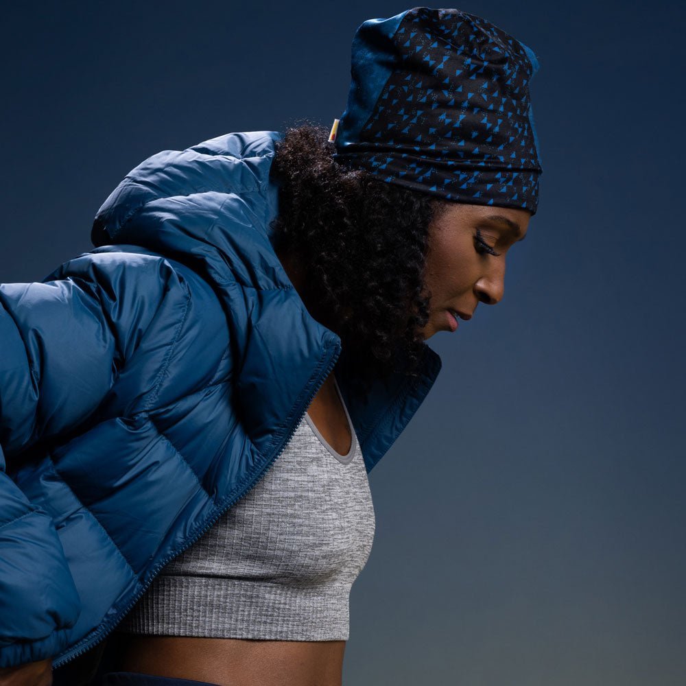 A woman wearing the Teal Flakes Beanie and a blue puffer jacket, looking down thoughtfully.