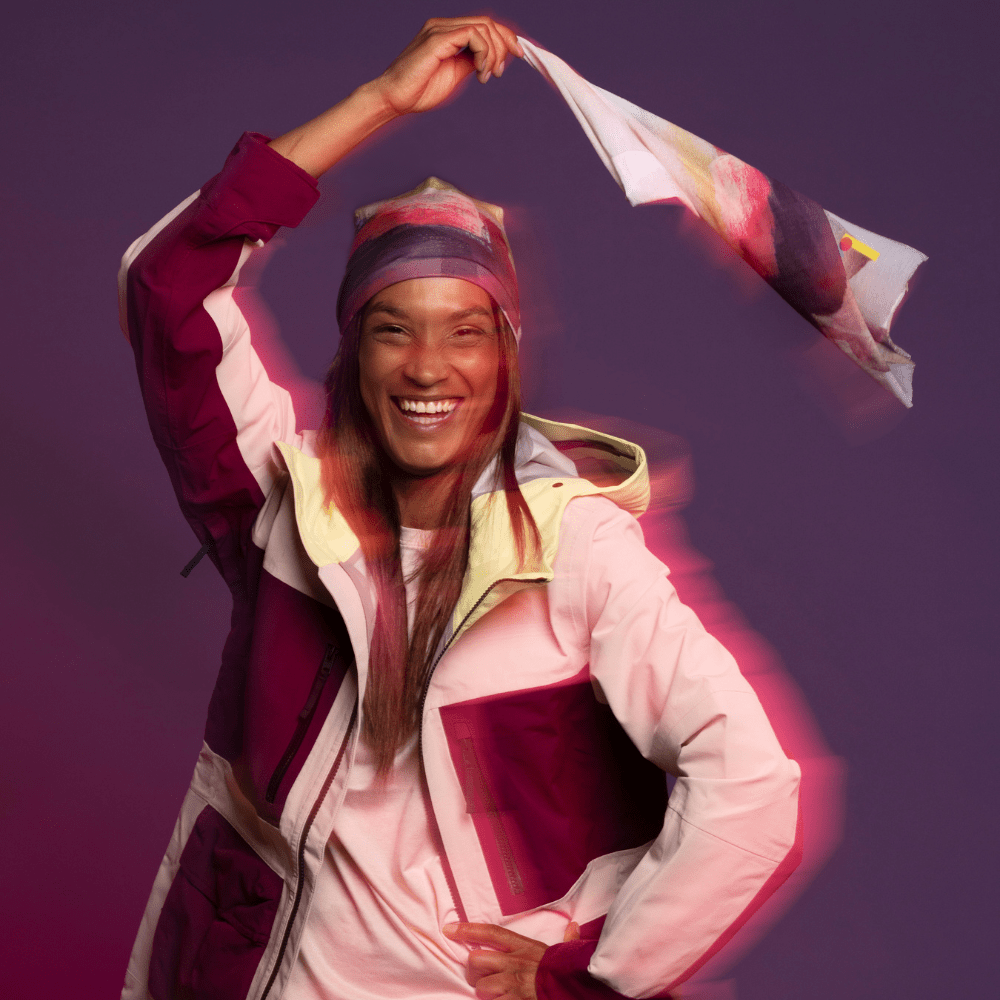 A smiling woman wearing The Space Between Beanie and a colourful ski jacket, holding up a matching scarf.