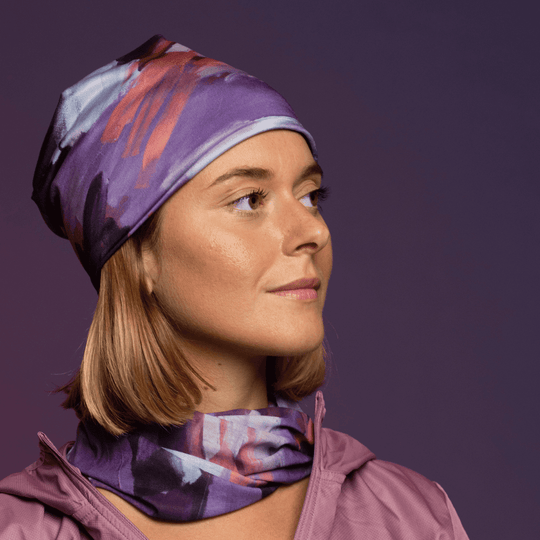 Woman wearing a purple abstract art pattern beanie and matching neck gaiter, looking thoughtfully to the side.