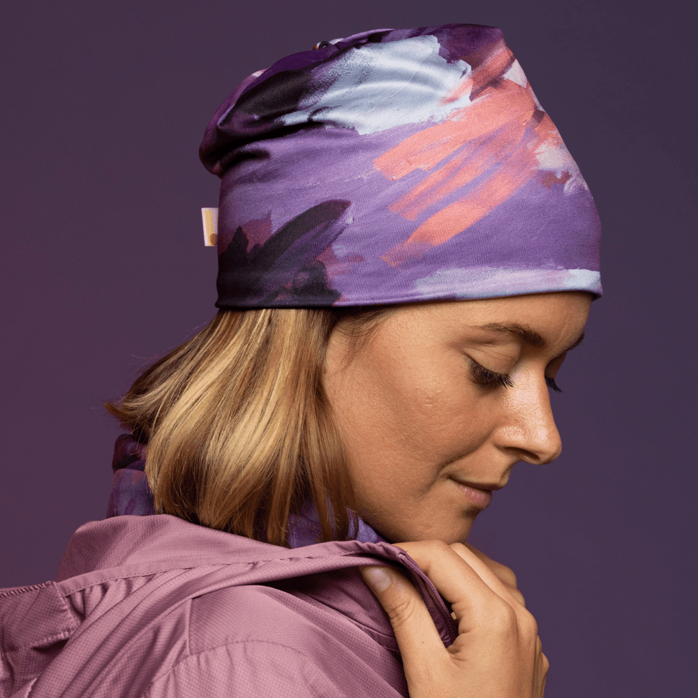 Woman wearing a purple abstract art pattern beanie and matching neck gaiter, with a pink jacket.