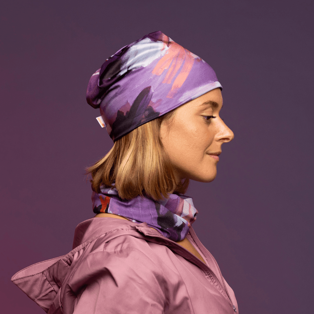 A woman in profile wears a matching beanie and neck tube with a floral art print, layered over a pink jacket.