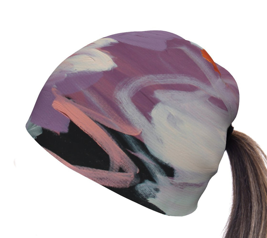 A purple and pink abstract art print covers a beanie designed for ponytails, with a swatch of hair emerging from the back opening.
