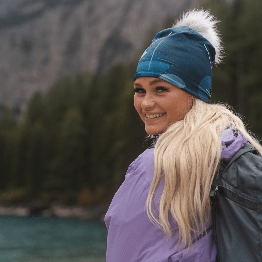 Smiling woman with blonde hair wearing the teal beanie with a grey pompom and a purple jacket, looking over her shoulder.