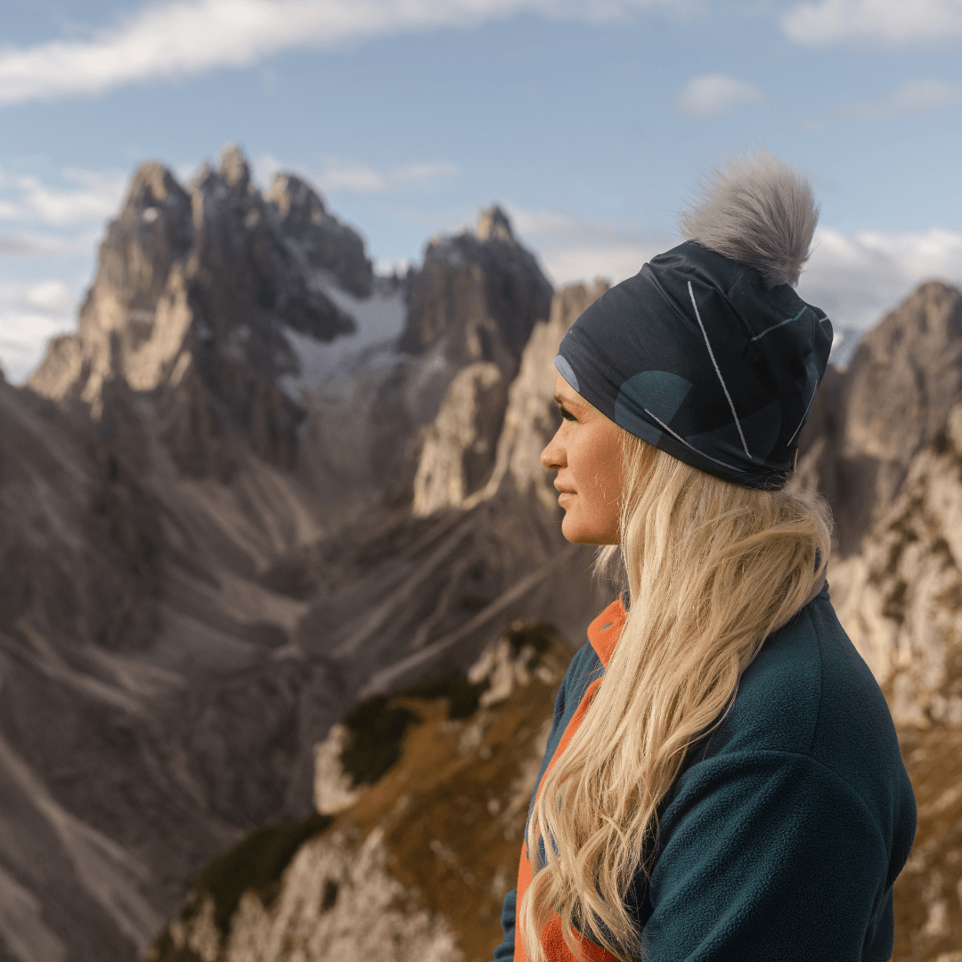 Woman wearing the teal beanie with a grey pompom while looking out at a mountainous landscape.