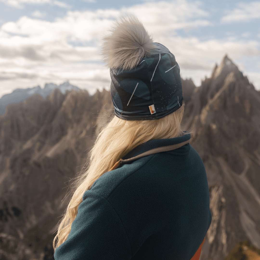 Back view of a woman wearing the teal beanie with a grey pompom in a mountain setting.
