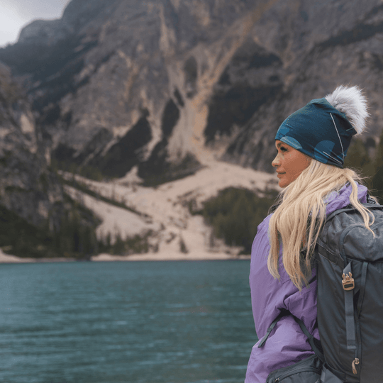 Woman wearing the teal beanie with a grey pompom and a purple jacket, looking out at a scenic lake with mountains in the background.