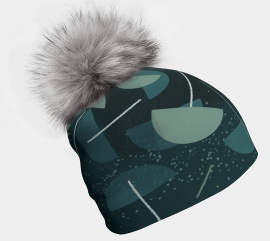 Close-up of a dark teal beanie with an abstract teal and green pattern and a fluffy grey pompom.