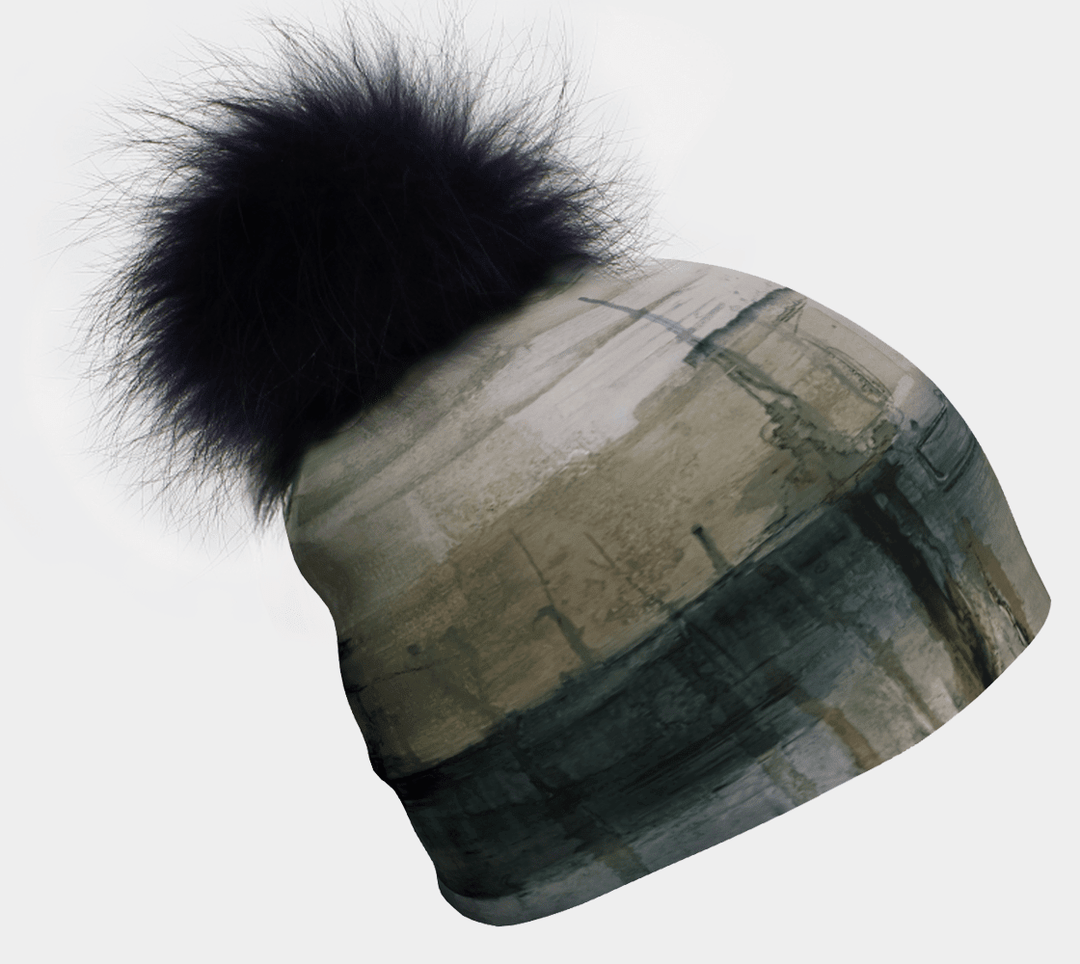 A green and grey abstract patterned beanie with a large black fur pompom is shown against a white background.