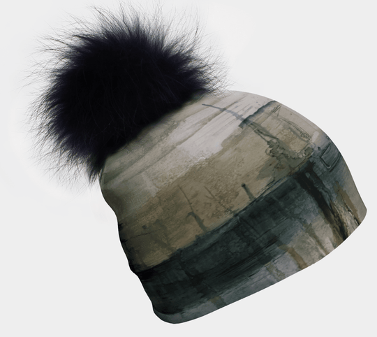A green and grey abstract patterned beanie with a large black fur pompom is shown against a white background.